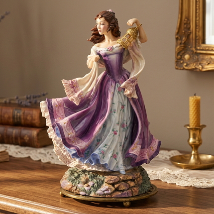 A decorative porcelain figurine of a woman in a flowing purple and pale blue gown, posed on a tabletop with books, lace, and a candle in the background.