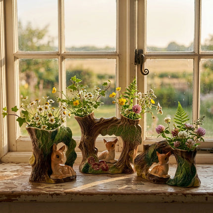 A ceramic woodland-themed planter or decorative piece featuring three stylized tree stumps with faux foliage and small painted deer figures, shown on a rustic wooden window sill.