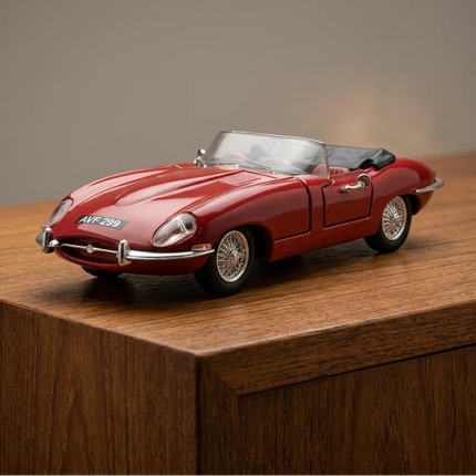 A red diecast model of a classic convertible sports car with a black soft top, photographed on a white surface with a branded patterned backdrop.
