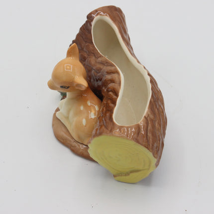 A ceramic decorative figurine shaped like a small deer tucked inside a log or tree stump, photographed on a plain white background.