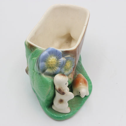 A ceramic planter shaped like a green leaf with a beige rectangular pot inside, decorated with blue flowers, a small white bunny, and a small orange shell on a plain white background.
