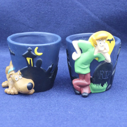 Two blue novelty shot glasses with Scooby-Doo themed decorations, one featuring Shaggy dog at a graveyard and the other featuring Fred Flintstone leaning on a tombstone, set against a blue background.