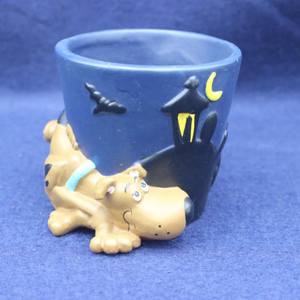 A blue ceramic pot featuring a sculpted character resembling Scooby-Doo lying on the front, with black bat and house details and a yellow crescent moon on the side, set against a blue background.