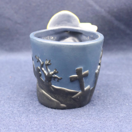 A small blue ceramic pot with a dark gray relief design of a cross and bare trees, with a white heart-shaped element visible at the top, set against a blue fabric background.