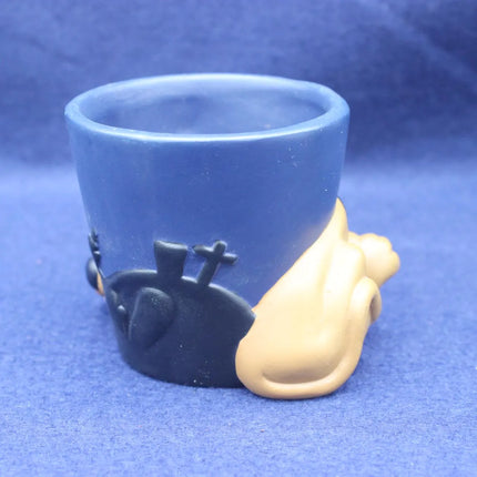A small blue cup or tumbler with sculpted black and beige decorative figures at the base, photographed against a blue fabric background.