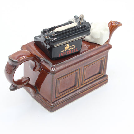 A ceramic teapot shaped like a brown wooden chest with a black typewriter and a white cat on top, featuring a glossy finish and the word 'IMPERIAL' printed on the typewriter.