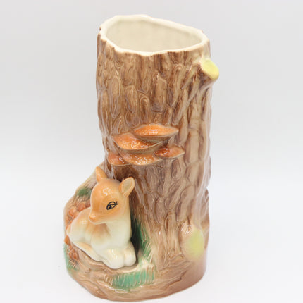 A ceramic woodland-themed vase shaped like a tree stump with a small deer figurine at the base, photographed against a plain light gray studio background.