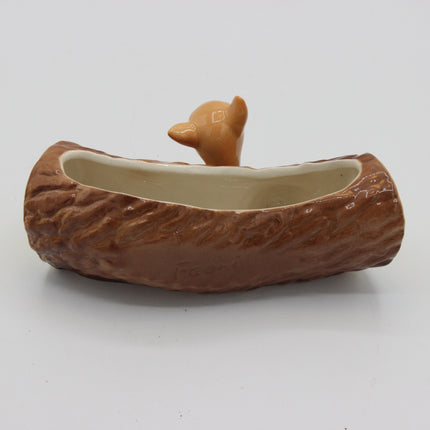 A ceramic planter shaped like a log cabin roof, with a small tan animal figure peeking out from the top and a shallow opening inside, photographed on a plain light background.