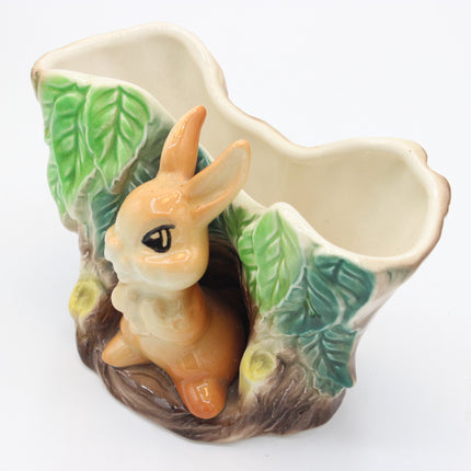 A ceramic vase designed as a forest scene, featuring a light brown rabbit sitting in a hollow, green leaves, and a brown tree trunk base, on a white background.