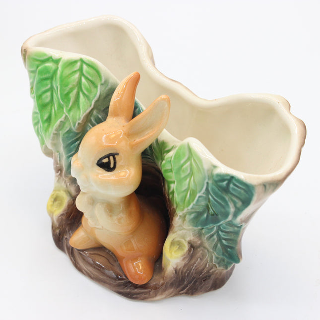 A ceramic vase designed as a forest scene, featuring a light brown rabbit sitting in a hollow, green leaves, and a brown tree trunk base, on a white background.