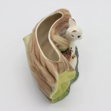 A ceramic animal figurine of a white cat with brown spots, nestled inside a stylized tree trunk or leaf-like shell with brown, beige, and green glaze details, on a plain white background.