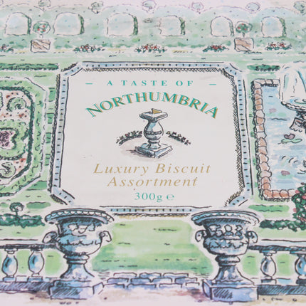 A close-up of a biscuit assortment package label reading 'A Taste of Northumbria Luxury Biscuit Assortment 300g e,' with an illustrated garden scene and urns in the background.