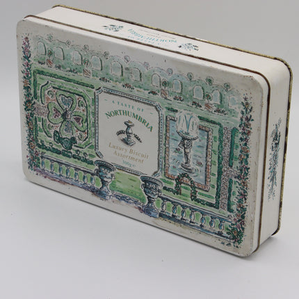 A vintage rectangular metal tin with rounded corners, featuring a printed landscape design in green, blue, and pink on an off-white background. The center label reads 'A Taste of NORTHUMBRIA Luxury Biscuit Assortment 1862'.