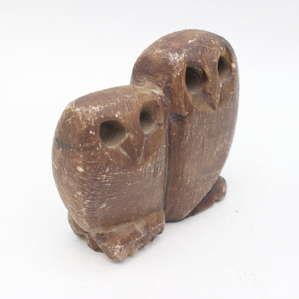A small carved stone owl figurine in a natural medium brown wood tone, featuring a larger owl standing behind a smaller owl, set against a plain white background.