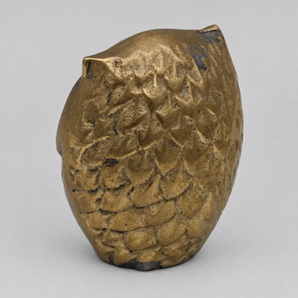 A small metallic gold owl figurine with a textured, scale-like surface, photographed on a plain light gray background.