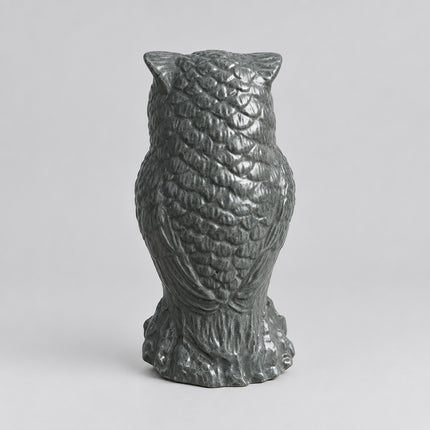 A small owl figurine with textured feather details, standing on a base, shown on a brown surface with a gray fabric background.