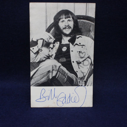 Black and white photo of Bill Oddie with long hair and a jacket, sitting on a chair.