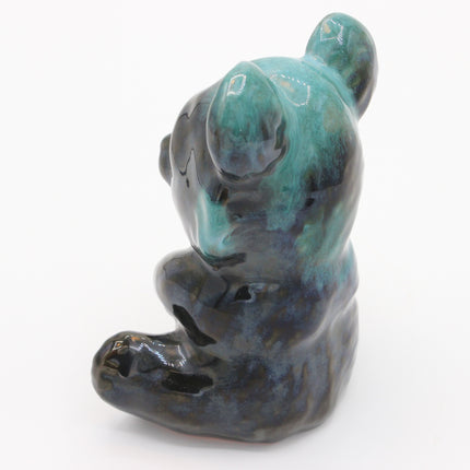 A small ceramic figurine of a seated bear, decorated with a glossy glaze featuring a mottled teal, blue, and dark charcoal color pattern, set against a plain white background.