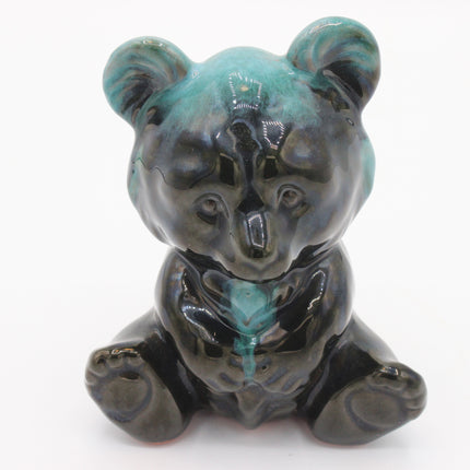 A ceramic figurine of a sitting bear with a glossy glaze, featuring a color-shifting effect that transitions from turquoise blue to dark gray and black. The figurine is photographed against a seamless white background.