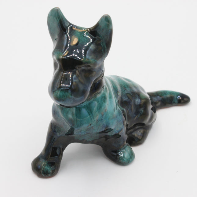 A glossy ceramic figurine of a crouching dog, featuring a dark teal and black glaze with white painted details on the face and chest, set against a plain white background.