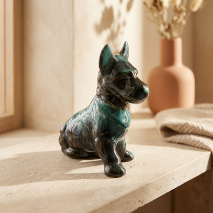 A glazed ceramic dog figurine in a sitting pose, featuring a dark green and black textured glaze with subtle blue-green highlights, displayed on a beige stone surface in a warm, sunlit interior setting.