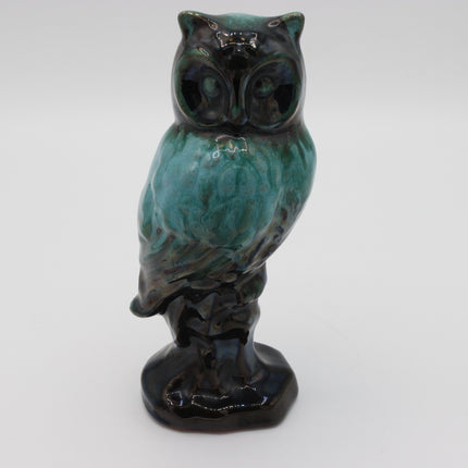 A ceramic owl figurine with a glossy teal and black glaze, standing on a dark base against a plain light gray background.