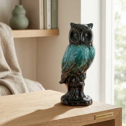 A ceramic owl figurine with a glossy teal and black glaze, displayed on a light wood table in a softly lit room.