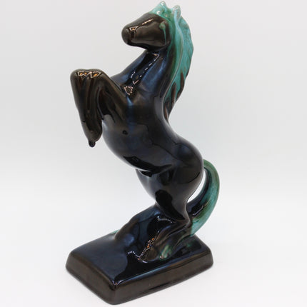 A glossy ceramic figurine of a rearing horse on a rectangular base, featuring a dark blue and black glaze with turquoise-green highlights on the mane, tail, and legs, set against a plain white background.