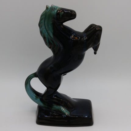 A glossy ceramic figurine of a rearing horse in a dark black glaze with vibrant teal-green accents on the mane, tail, and lower body, set against a neutral light gray background.