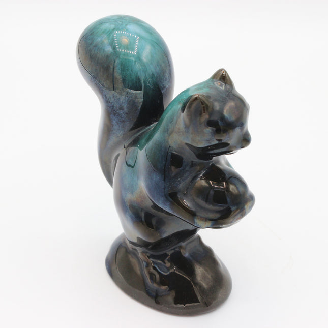 A glossy ceramic squirrel figurine holding a nut, featuring a dark blue and black glaze with teal-green highlights, set against a plain white background.
