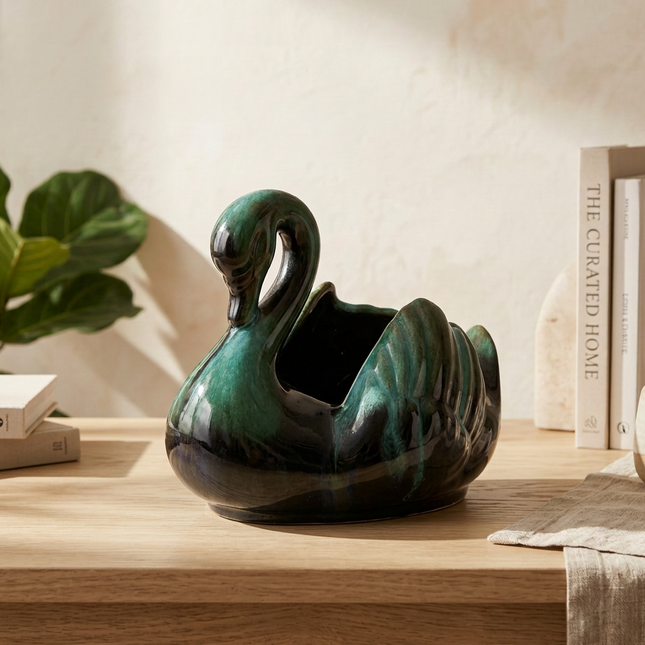 A glossy ceramic swan-shaped planter in deep green and black sits on a light wood table in a styled interior setting with books, a plant, and a draped fabric runner.