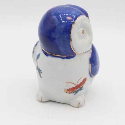 A small ceramic owl figurine with a glossy blue and white glaze, decorated with gold trim and painted details of a red butterfly and blue leaves, set against a plain white background.