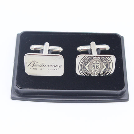 A pair of silver-toned Budweiser cufflinks displayed on a black presentation box. One cufflink is a simple rectangular plate engraved with 'Budweiser KING OF BEERS', and the other is a more detailed oval emblem featuring the number 25.