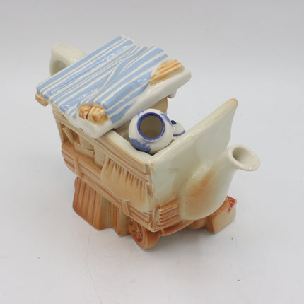A vintage ceramic teapot shaped like a small carriage or cart, featuring a cream-colored body, a blue and white striped roof, and a light brown wood-grain base, photographed on a neutral light gray background.