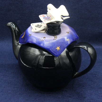 Black teapot with decorative lid featuring playing cards and a dove on a blue background