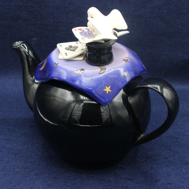 Black teapot with decorative lid featuring playing cards and a dove on a blue background