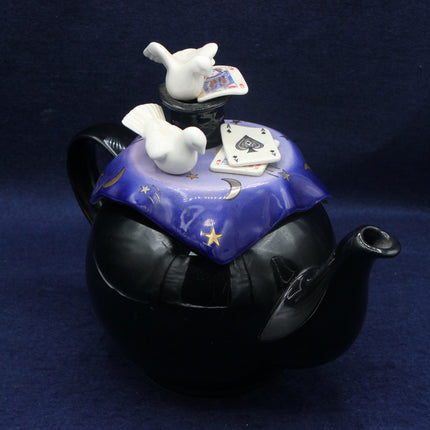 Black teapot with a blue lid featuring white doves and playing cards on a dark background