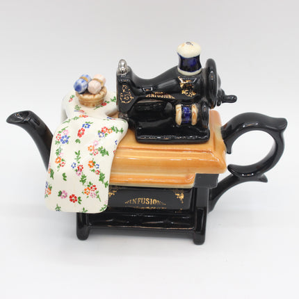 A decorative ceramic teapot shaped like a sewing machine, featuring a black body with gold lettering, a peach-colored top panel, and a draped cream fabric with a colorful floral print.