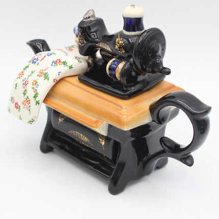 A ceramic sewing machine figurine with a black body, gold accents, a light wood-grain tabletop, and a white floral fabric roll, photographed on a white background.