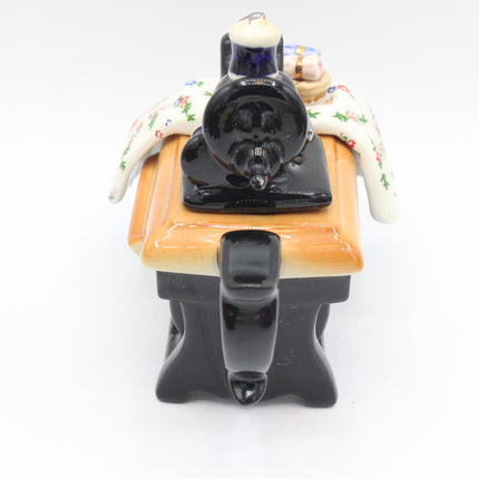 A ceramic table-top figurine depicting a black steam locomotive pulling a passenger car with a patterned cream tablecloth, set against a plain white background.