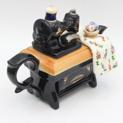 A decorative ceramic figurine of a black sewing machine with gold detailing, a light wood-grain tabletop, and a cream floral cloth draped over the side, set against a plain white background.