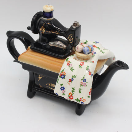 A decorative ceramic sewing machine teapot on a white background. The teapot features a black body, a light wood-grain top, a floral cream-colored fabric draped over the side, and a small ceramic spool of thread on top.
