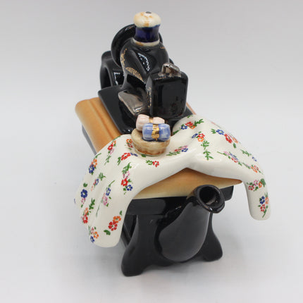 A ceramic novelty figurine shaped like a black teapot or small vessel with a figure in dark clothing holding a small object, draped with a white tablecloth printed with small colorful flowers, on a light wood-toned base against a plain white studio background.