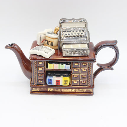 A decorative ceramic teapot shaped like a miniature tea shop counter, featuring a brown body with gold detailing, a silver cash register on top, and colorful tea bag and book accessories.