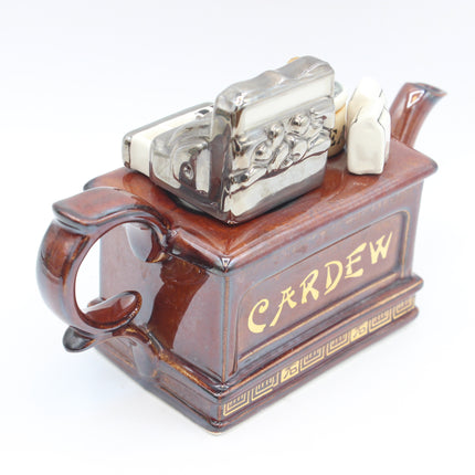 A ceramic tea towel holder shaped like a brown teapot, featuring the word 'CARDEW' in gold lettering on the front. The top is decorated with a silver-toned metal lion emblem and small white ceramic figures, set against a plain white background.