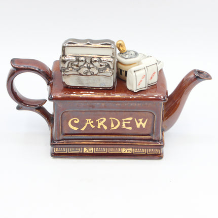 A ceramic teapot-shaped container with the name 'CARDEW' in gold lettering on the front, featuring a brown glazed body, a small silver ornate box on top, and a small cream-colored tag with a gold loop, photographed against a plain white background.