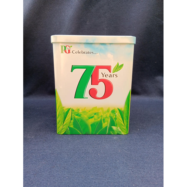 A square tin container celebrating 75 years, featuring a white top half with a blue sky graphic and green leaves, and a green leafy patterned bottom half. The front displays 'DG Celebrates.