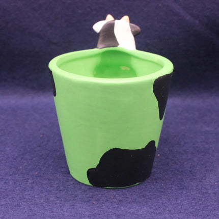 A green ceramic cup or candle holder with black cow-print spots and a small black-and-white cow figurine peeking out from the top, set against a dark purple background.