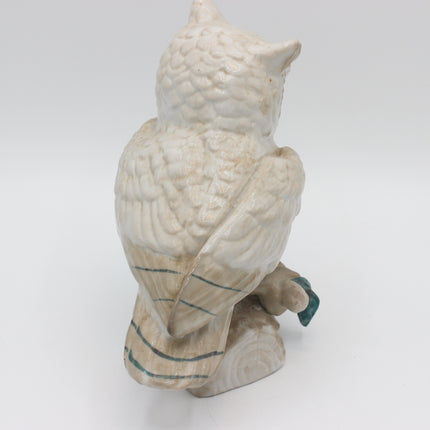 An off-white ceramic owl figurine shown from the back, perched on a small tree stump base with a small teal accent on its foot.