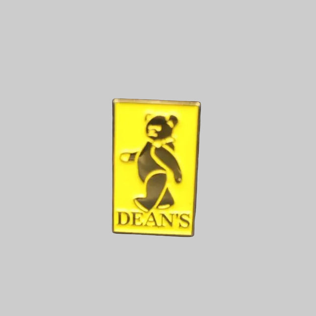 A small yellow rectangular enamel pin featuring a black teddy bear and the text 'DEAN'S' in black, placed on a dark blue textured fabric background.